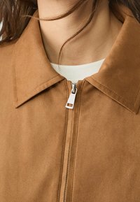 Chaqueta de ante marrón con un cuello afilado y cremallera plateada. La tela tiene una textura suave con un diseño limpio y minimalista.