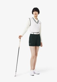 Weißer ärmelloser Pullover mit Rautenmuster über einem Langarmshirt, kombiniert mit dunkelgrünen Shorts und weißen Sport Schuhen. Golfclub in der Hand.