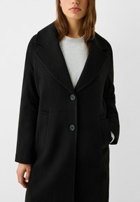 Svart ullkappa med stor krage, två framfickor och två knappar. Bärs över en grå tröja och visar upp en strukturerad design.