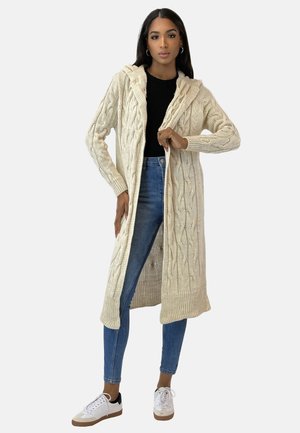 Langer, cremfärbiger Cardigan aus Zopfmuster mit Kapuze, offener Vorderseite, gerippten Bündchen und Saum, getragen über einem schwarzen Oberteil und blauen Jeans mit weißen Sneakern.