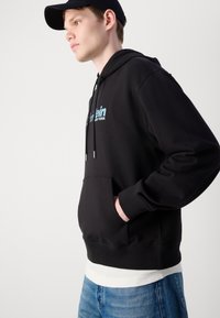 Black hooded sweatshirt with front pocket, ribbed cuffs, and hem. Features blue logo text. Worn with light blue denim jeans.