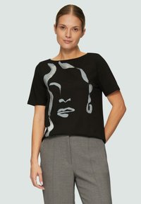 Zwart T-shirt met korte mouwen en een zilveren abstract gezicht ontwerp. Zachte stof, losse pasvorm, ronde halslijn, gecombineerd met grijze broek.