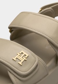 Κοντινό πλάνο λουριών μπεζ σανδαλιού με χρυσούς διασταυρούμενους λογότυπους TH σε ρυθμιζόμενο κούμπωμα, που δείχνει ομαλή υφή και λεπτομέρειες ραφής.