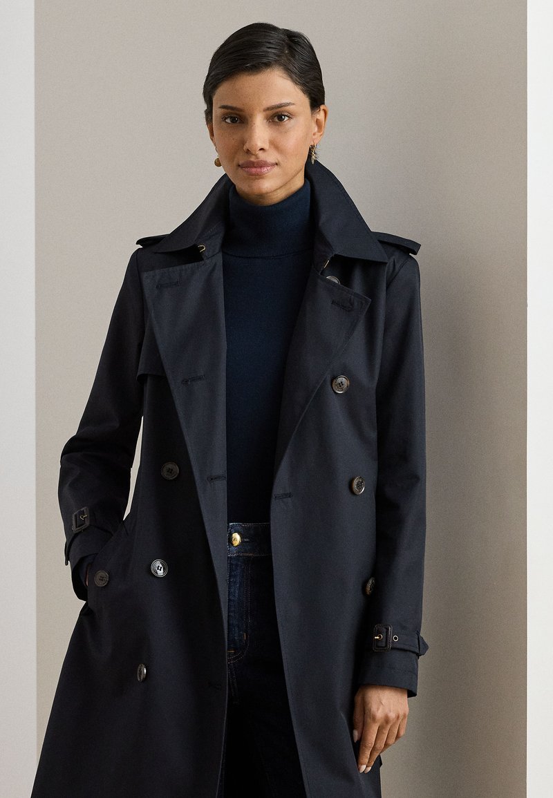 Trench-coat bleu marine en tissu lisse, avec un design croisé à double boutonnage, de gros boutons, des épaulettes et des revers prononcés.