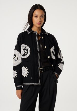 Zwarte stoffen jas met ingewikkelde witte bloemenborduursels, een knoopsluiting aan de voorkant en contrasterende stiksels langs de kraag en manchetten.