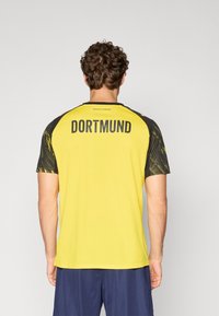 Gul atletisk skjorte med korte sorte mønstrede ærmer, med "DORTMUND" trykt på ryggen. Struktureret stof, rund hals-design.