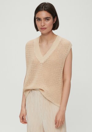 Gilet en tricot beige avec un décolleté en V et des manches courtes, présentant un motif de trame ajourée. Porté sur un pantalon beige léger et plissé.