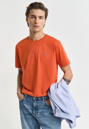 Giovane uomo con capelli castani corti che indossa una maglietta arancione e jeans blu, con una camicia celeste a maniche lunghe piegata su un braccio.