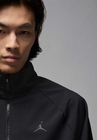 Chaqueta negra con cremallera y cuello alto, fabricada con un tejido suave. Presenta un logo Jumpman gris en el lado izquierdo, enfatizando un diseño atlético.