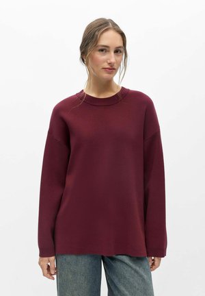 Ung kvinde med lyst brunt hår iført en løs vinrød sweater og blå jeans, stående mod en ensfarvet hvid baggrund.