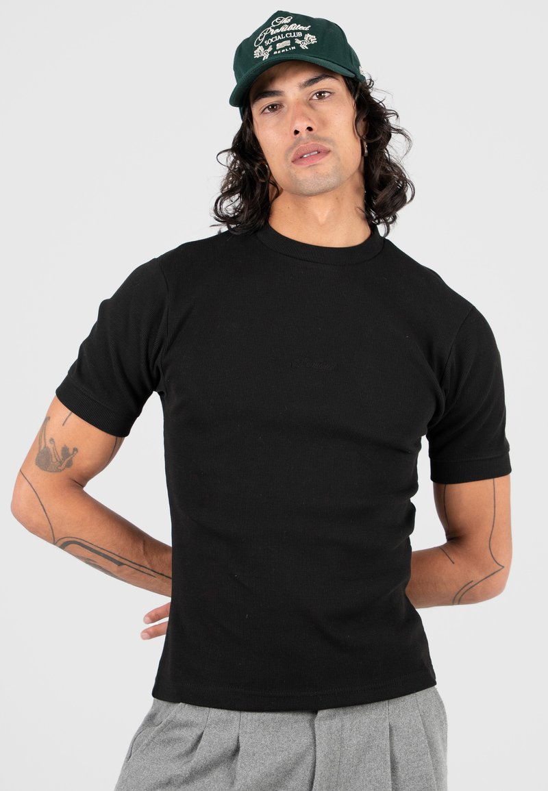 Uomo con capelli lunghi che indossa una maglietta nera, pantaloni grigi, un berretto verde e tatuaggi sulle braccia, posa con le mani dietro la schiena su uno sfondo bianco.