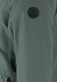 Grön vattentät jacka med en slät textur och en cirkulär svart logotyp patch på överarmen, med kontrasterande sömnadsdetaljer.