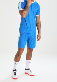 Camiseta atlética azul con acentos blancos y pantalones cortos a juego, hechos de material ligero. Calcetines blancos y zapatos negros con suelas rojas.