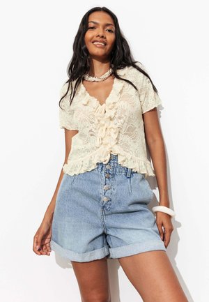 Femme portant un chemisier en dentelle crème à manches courtes avec des volants et un short en jean bleu clair taille haute boutonné.