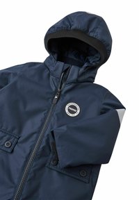 Marineblauwe waterdichte jas met capuchon, ritssluiting aan de voorkant, twee opgestikte zakken en een rond logo patch. Bevat grijze accenten op de mouwen.