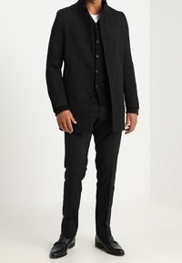 Cappotto nero su misura con un motivo testurizzato, chiusura a bottoni e colletto alzato, abbinato a un gilet e pantaloni neri, completato da scarpe nere.