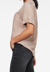 Camicia beige chiaro a maniche corte in un tessuto morbido e leggermente lucido; presenta maniche arrotolate e un'etichetta discreta sul lato, abbinata a jeans scuri.