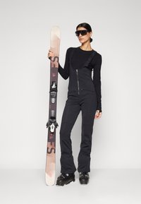 Svart skianlegg med glidelåser og lommer, kombinert med ski og svarte støvler. Tilbehør inkluderer overdimensjonerte solbriller.