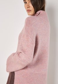 Rosa Strickpullover mit hohem Rippkragen, lockerer Passform, seitlichen Schlitzen und strukturiertem Garn. Weiches, gemütliches Material mit subtilen Farbvariationen.