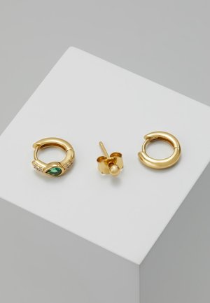 Dwa złote kolczyki koła, jeden z zielonym kamieniem i małymi przezroczystymi kamieniami, oraz jeden prosty złoty kolczyk na białej powierzchni.