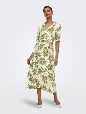 Žena stojaca v béžových midi šatách so zelenými vzormi listov, krátkymi rukávmi, golierom, previazaným pásom a bielymi lodičkami s bodcom na špičke.