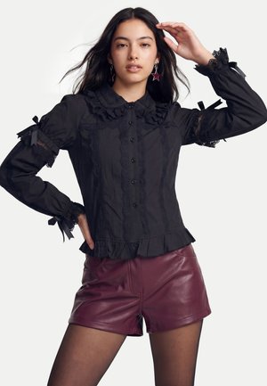 Ung kvinde iført sort blondebluse og bordeaux lædershorts poserer med den ene hånd på hovedet og den anden på hoften mod hvid baggrund.