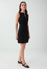 Woman standing wearing a sleeveless black dress with a small front keyhole and black Mary Jane flats against a plain light background.