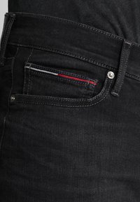 Jeans de mezclilla negra con un ajuste slim, que presentan un borde de bolsillo contrastado en azul, blanco y rojo, y un cierre de botón plateado.