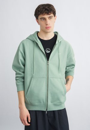 Joven con cabello corto y rizado, vistiendo una sudadera con capucha verde claro con cremallera sobre una camiseta negra de Nike, de pie contra un fondo claro y liso.