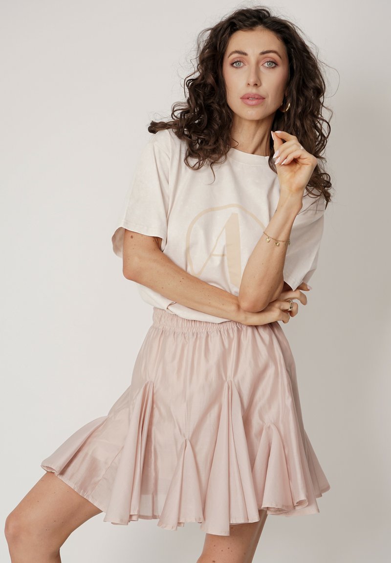 Jasnoróżowa rozkloszowana spódnica z warstwowymi falbanami i elastycznym paskiem w talii, zestawiona z kremowym t-shirtem z okrągłym logo.