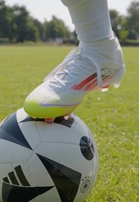 Chaussure de football blanche avec des accents rouges et un bout vert, positionnée sur un ballon de football à motif noir et blanc sur de l'herbe.