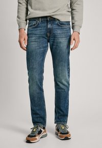 Tmavomodré džínsy so slim fit strihom, s piatimi vreckami, svetlým opotrebovaním a jemnými švovými detailmi. Nosia sa s viacfarebnými teniskami.