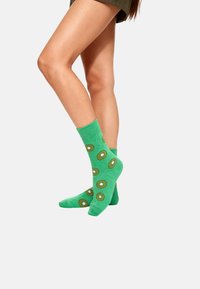 Piernas que llevan calcetas verde hasta la mitad de la pantorrilla con patrones de rodajas de kiwi sobre un fondo blanco.