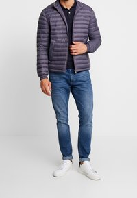 Giubbotto piumino di colore grigio scuro con motivo a righe orizzontali, collo alto, chiusura a zip e polsini a costine, abbinato a jeans blu e sneakers bianche.