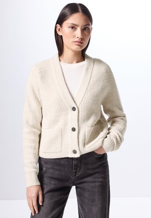 Cremfarbener, gerippter Cardigan mit Schalkragen, fünf Knöpfen und zwei vorderen Taschen, kombiniert mit dunklen Jeans. Glatte Textur, lässige Passform.