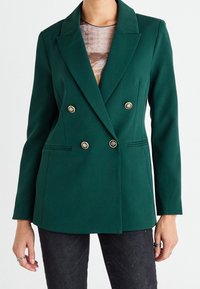 Femme portant un blazer croisé vert foncé à boutons dorés sur un haut à motifs et un jean sombre, les mains détendues le long du corps.
