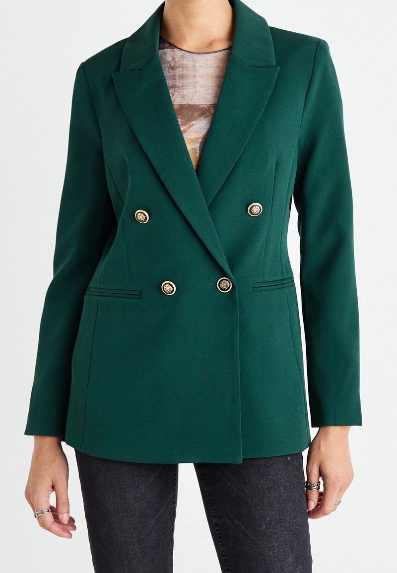 Femme portant un blazer croisé vert foncé à boutons dorés sur un haut à motifs et un jean sombre, les mains détendues le long du corps.