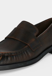 Σκούρο καφέ δερμάτινο μοκασίνι με ραφές μοκ τού, φαρδιά λωρίδα στο επάνω μέρος και μαύρη καουτσούκ σόλα.