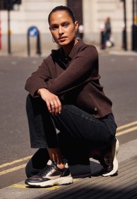 Brązowy sweter z kołnierzykiem, czarne dżinsy z detalem na guziki oraz czarno-kremowe sneakersy z siateczkowymi akcentami. Miejski krajobraz w tle.