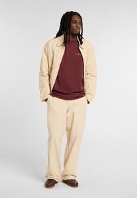 Beige bavlněná bunda s límečkem, vínové tričko se znakem, béžové kalhoty a tmavě hnědé boty. Celkově hladká textura.