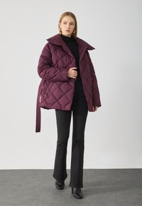Giacca trapuntata bordeaux con ampio colletto, bottoni anteriori e cintura, abbinata a pantaloni neri a campana e stivaletti.