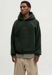 Sweat à capuche vert en polaire avec une surface textur�ée et pelucheuse, comportant une poche avant et des poignets côtelés, porté par-dessus une chemise à carreaux et un pantalon beige.