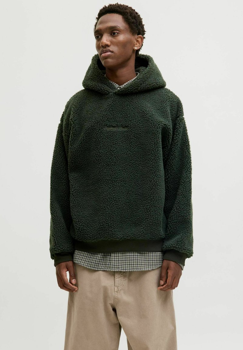 Sweat à capuche vert en polaire avec une surface texturée et pelucheuse, comportant une poche avant et des poignets côtelés, porté par-dessus une chemise à carreaux et un pantalon beige.