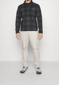 Homme portant un pull polaire à fermeture éclair quart en damier noir et gris, un pantalon cargo beige et des chaussures de sport grises, blanches et orange, debout sur un fond uni.