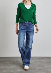 Grüner, langärmeliger V-Ausschnitt-Pullover über einem weißen T-Shirt, kombiniert mit lockeren blauen Jeans und weißen Sneakers. Glatte Texturen.