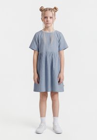 Mergaitė stovinti atsisukusi į priekį, dėvinti mėlyną dryžuotą trumpomis rankovėmis suknelę, baltas kojines ir baltus sportbačius, plaukus surišusi dviem pintinėmis kuodeliais.