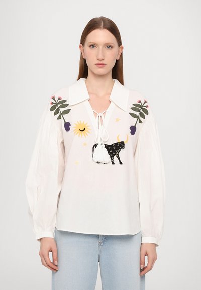 Blouse blanche avec un large col, ornée de broderies florales colorées et de soleils, d'un graphique de taureau noir et d'accents à franges. Manches longues et bouffantes.