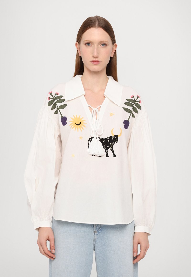 Weiße Bluse mit weitem Kragen, verziert mit bunten Blumen- und Sonnenstickereien, einem schwarzen Stiergrafik und Quastenakzenten. Lange, voluminöse Ärmel.