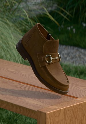 Bottine en suède marron avec un bout arrondi, une fermeture éclair latérale et un détail en mors doré, posée sur un fond naturel.