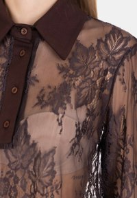 Donna che indossa una blusa in pizzo nero trasparente con motivi floreali e un colletto e bottoni neri solidi sopra un reggiseno beige.